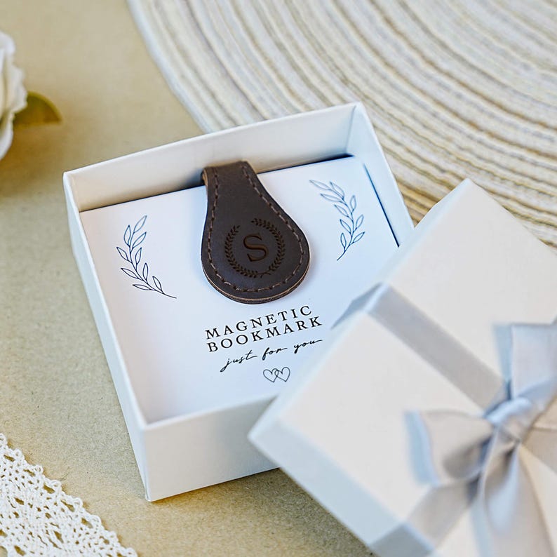 Pu&ograve; includere: Un segnalibro magnetico in pelle marrone con la lettera "S" in una ghirlanda decorativa, all'interno di una confezione regalo bianca. La scatola riporta la scritta "MAGNETIC BOOKMARK just for you". La scatola &egrave; legata con un nastro grigio.