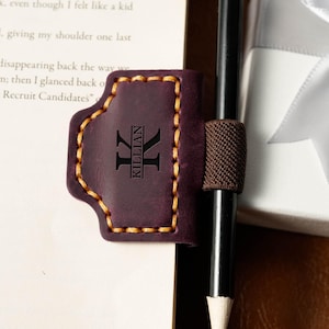 May include: A dark purple leather pen holder with the name "KILLIAN" and a stylized "K" engraved on it. The holder has a brown elastic band and is stitched with yellow thread. A black pencil is held in place.