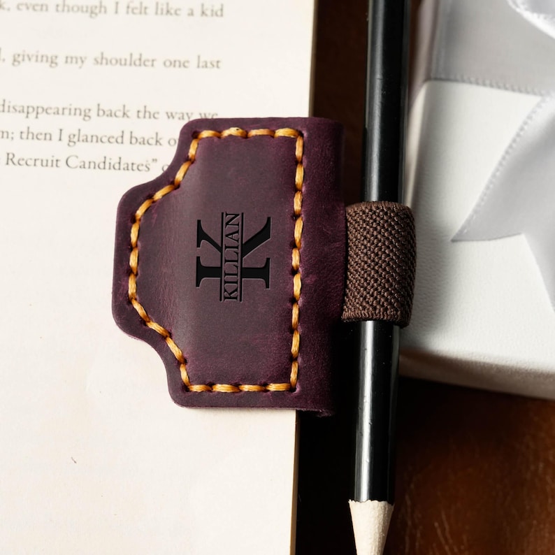 May include: A dark purple leather pen holder with the name "KILLIAN" and a stylized "K" engraved on it. The holder has a brown elastic band and is stitched with yellow thread. A black pencil is held in place.