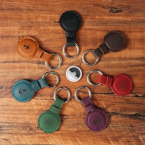 May include: A collection of leather keychains in various colors, including black, brown, red, green, teal, tan, and purple. Each keychain features a circular leather holder with a silver keyring. Some holders have embossed designs or initials.