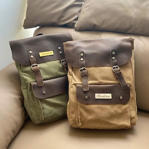 May include: Two canvas backpacks with leather accents. The green backpack has the name "Andrew" on it. The brown backpack has the name "Christine" on it.
