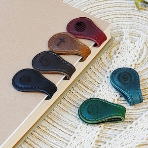 Pu&ograve; includere: Una collezione di segnalibri magnetici in pelle in vari colori, tra cui nero, marrone e bordeaux. Ogni segnalibro presenta un design unico, come iniziali, una croce o una bandiera americana. I segnalibri sono esposti su un libro.