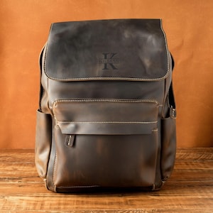 May include: A dark brown leather backpack featuring a large top flap with the engraved name KILLIAN centered on the front. The bag includes a spacious front pocket with a horizontal flap closure, side pockets, and visible light-colored stitching.