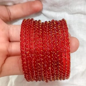 May include: A collection of red glass bangles with a textured finish and tiny gold-coloured embellishments. The bangles are stacked, highlighting their detailed design and rich colour. These accessories are ideal for adding a touch of elegance to any outfit.