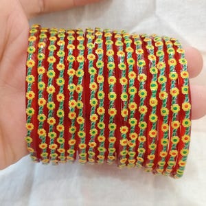 Può includere: Una pila di braccialetti rossi con un motivo floreale. I braccialetti sono adornati con fiori gialli e verdi, creando un motivo vibrante e decorativo. I braccialetti sono tenuti in una mano.