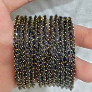 以下が含まれることがあります： ゴールドのアクセントが付いた黒いバングルのセット。バングルは積み重ねられており、小さく均等に配置された金色のビーズが付いたテクスチャードデザインを示しています。ジュエリーはニュートラルな背景に表示されます。