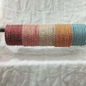 Puede incluir: Un conjunto de pulseras coloridas con pedrería. Las pulseras están dispuestas en fila y están hechas de un material brillante. Las pulseras son en tonos de rojo, rosa, blanco, dorado y azul.