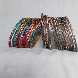 Puede incluir: Una pila de pulseras de vidrio de colores con pedrería. Las pulseras son en tonos de azul, verde, rosa y naranja. También hay algunas pulseras plateadas con pedrería.