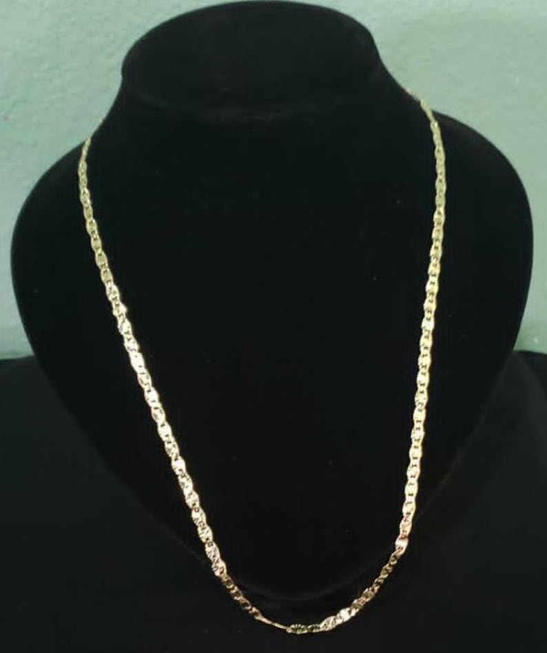 May include: A gold-colored chain necklace with a twisted link design. The necklace is displayed on a black mannequin bust. The chain has a shiny finish and is suitable for everyday wear or special occasions.