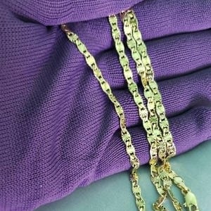 May include: A gold-colored necklace with a repeating starburst pattern. The chain is displayed on a purple fabric, showcasing the intricate design and the necklace's shine. The necklace is a jewelry item.