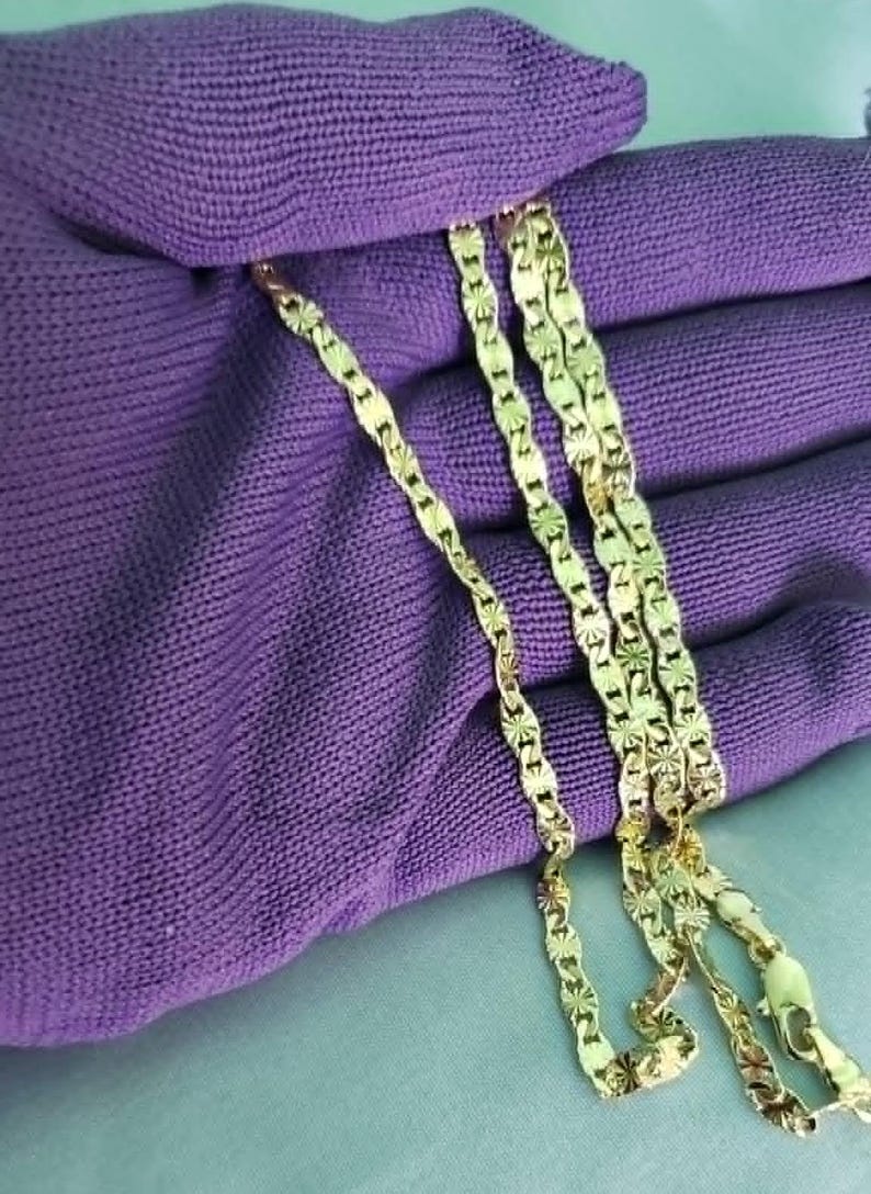 May include: A gold-colored necklace with a repeating starburst pattern. The chain is displayed on a purple fabric, showcasing the intricate design and the necklace's shine. The necklace is a jewelry item.
