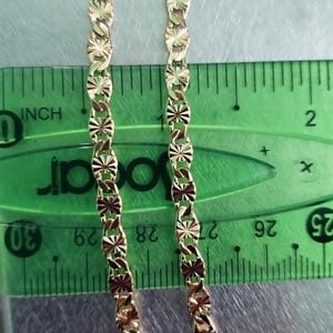 May include: Two gold-colored necklaces with a repeating starburst design are displayed against a green ruler. The ruler shows measurements in inches. The necklaces are approximately 1 inch wide.
