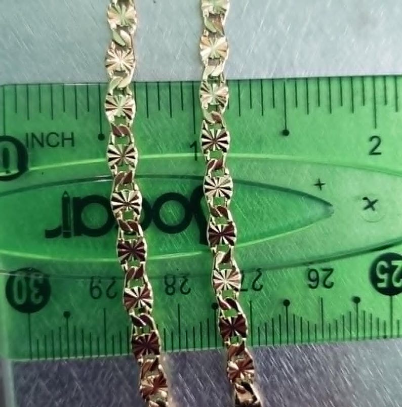May include: Two gold-colored necklaces with a repeating starburst design are displayed against a green ruler. The ruler shows measurements in inches. The necklaces are approximately 1 inch wide.