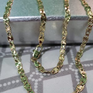 May include: A gold-colored necklace with a delicate, starburst-shaped link design. The chain is displayed against a soft, neutral background, highlighting its intricate pattern and the warm tone of the metal. The necklace is secured with a small clasp.