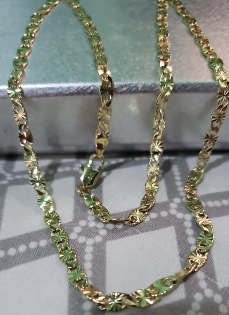 May include: A gold-colored necklace with a delicate, starburst-shaped link design. The chain is displayed against a soft, neutral background, highlighting its intricate pattern and the warm tone of the metal. The necklace is secured with a small clasp.