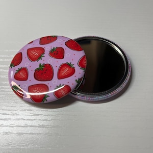 May include: A compact mirror with a pink lid featuring a pattern of red strawberries with white stars. The mirror is open and the reflective surface is visible.