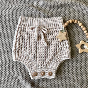 Peut inclure: Un bloomer de bébé tricoté beige avec un nœud coulissant et trois boutons en bois. Une attache-tétine en forme d'étoile en bois est attachée à une chaîne de perles en bois. Les articles sont sur une couverture texturée de couleur neutre.