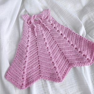 May include: A pink crocheted baby top with a delicate openwork pattern. The top has a halter neckline and a loose, flowing fit.