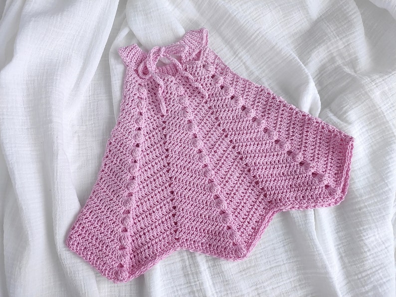 May include: A pink crocheted baby top with a delicate openwork pattern. The top has a halter neckline and a loose, flowing fit.