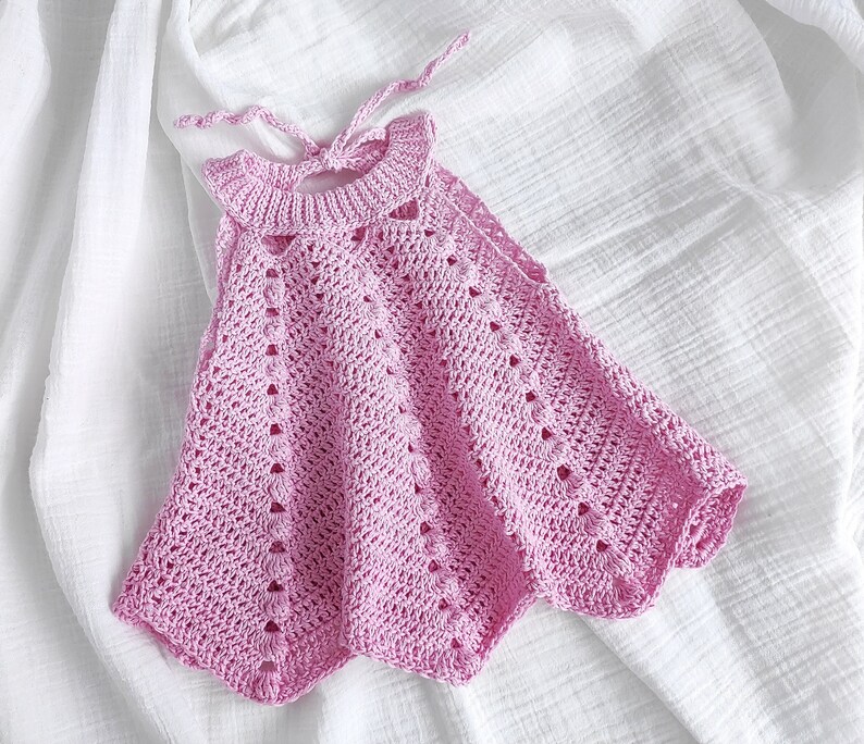 May include: A pink crocheted sleeveless dress with a delicate openwork pattern. The dress has a halter neckline and a flowing skirt.