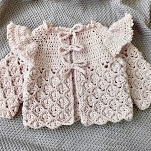 Può includere: Un cardigan per neonato in crochet rosa con scollo a volant e maniche lunghe. Il cardigan ha una chiusura a laccio sul davanti.