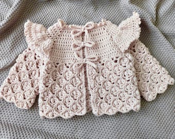 May include: A pink crocheted baby cardigan with a ruffled neckline and long sleeves. The cardigan has a tie closure at the front.