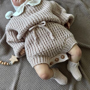 Puede incluir: Muñeca bebé con un conjunto de punto marrón claro, que incluye un suéter, pantalones cortos y botines. El conjunto tiene un patrón texturizado y botones de madera. También hay un mordedor en forma de estrella de madera con cuentas.