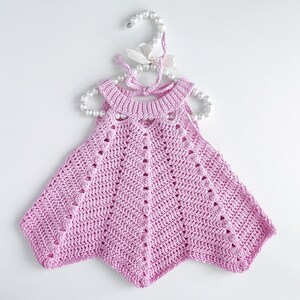May include: A pink crocheted baby dress with a scalloped hem and a delicate openwork design. The dress is hanging on a white hanger with pearl beads.