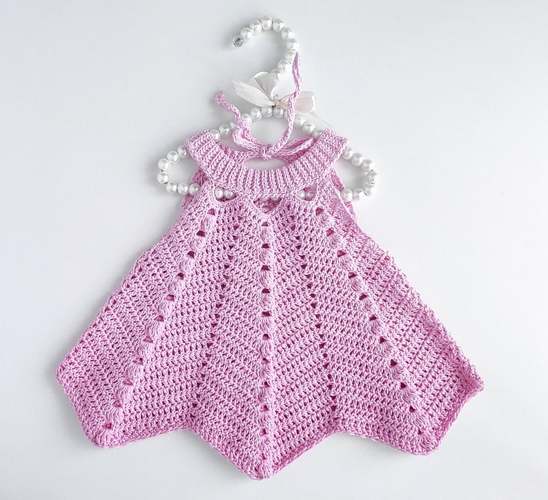May include: A pink crocheted baby dress with a scalloped hem and a delicate openwork design. The dress is hanging on a white hanger with pearl beads.