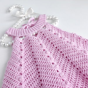 May include: A pink crocheted baby dress with a delicate openwork pattern. The dress is laid out on a white surface with a pearl necklace draped over it.