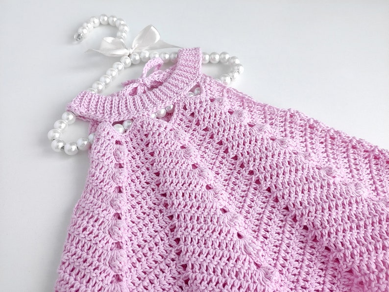 May include: A pink crocheted baby dress with a delicate openwork pattern. The dress is laid out on a white surface with a pearl necklace draped over it.