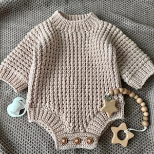 Peut inclure: Un body pour bébé en tricot marron clair avec des manches longues et un col côtelé. Le body a trois boutons en bois en bas et est accompagné d'une sucette et d'un anneau de dentition en bois avec des perles et des étoiles.