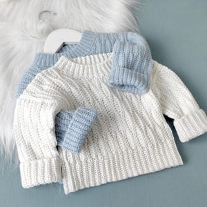Può includere: Un maglione bianco lavorato a uncinetto con maniche lunghe e collo rotondo. Il maglione è abbinato a un cappello lavorato a uncinetto blu chiaro e guanti abbinati.