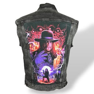 May include: A black denim vest featuring a wrestler graphic with a hat, purple gloves, and a gold container. The back of the vest has a graveyard scene with a full moon and red-eyed demons in the background.