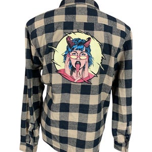 May include: A tan and navy blue plaid flannel shirt with a graphic on the back. The graphic is a cartoon image of a woman with blue hair, devil horns, and glasses, sticking out her tongue.