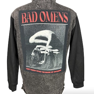 Könnte beinhalten: Eine schwarze und graue Jeansjacke mit schwarzen Ärmeln. Auf der Rückseite der Jacke befindet sich eine Grafik mit den roten Worten "BAD OMENS" und ein Schwarz-Weiß-Bild einer Person. Der Text "I DON'T KNOW WHAT I'M MADE OF ANYMORE" steht darunter.