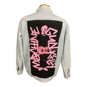 May include: Light blue denim jacket with a black back panel featuring a pink graphic of a person holding a guitar, with the words "MACHINE GUN KELLY" in pink. The jacket has a classic collar and button closures.