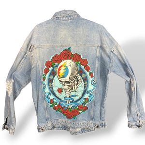 May include: Light blue denim jacket with a large graphic on the back featuring a skull, roses, and a lightning bolt design. The jacket has a classic collar, button closures, and distressed details on the sleeves.