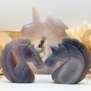 May include: Three carved stone unicorn figurines. Two are in the foreground, facing each other, with shades of gray and purple. The third, larger unicorn is in the background, in a light pink hue. The background includes dried flowers.