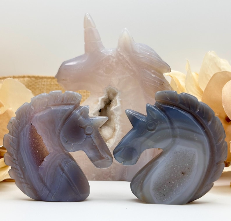 May include: Three carved stone unicorn figurines. Two are in the foreground, facing each other, with shades of gray and purple. The third, larger unicorn is in the background, in a light pink hue. The background includes dried flowers.