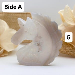 May include: A carved, light gray unicorn figurine. The sculpture is made of a translucent stone with subtle variations in color and texture. The unicorn's mane and horn are detailed. The text "Side A" is in a white box in the upper left corner.