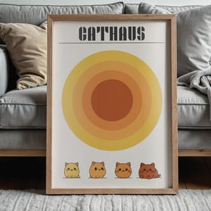 Op de afbeelding: Een ingelijste print met het woord "CATHAUS" boven een retro zonne-ontwerp in geel en oranje. Daaronder staan vier cartoonkatten in tinten oranje en geel. De print wordt in een woonkamer getoond, wat een vleugje kleur toevoegt.