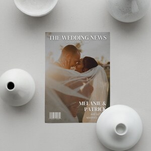 May include: A magazine cover titled "THE WEDDING NEWS" featuring a couple embracing. The cover includes the date "SATURDAY, AUGUST 15, 2024" and the names "MELANIE & PATRICK". White vases surround the magazine on a gray surface.
