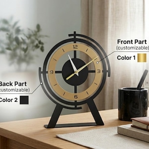 Puede incluir: Un reloj de escritorio negro y dorado con marco circular y soporte. La esfera presenta un centro negro, un anillo dorado con los números 3, 6, 9 y 12, y manecillas doradas. Etiquetas de texto indican partes personalizables con opciones de color.