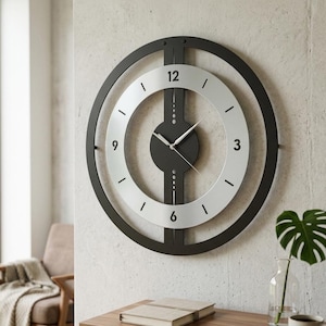 May include: A circular wall clock featuring a black outer frame and a silver inner ring with black numerals 3, 6, 9, and 12. A vertical black bar runs through the center, connecting to a dark circular hub with thin silver hour and minute hands.