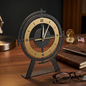 May include: A circular desk clock featuring a dark gray textured frame and a gold-colored inner ring with black numerals at the 12, 3, 6, and 9 positions. The clock has thin gold hands and a central dark gray circular accent piece.