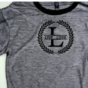 K&ouml;nnte beinhalten: Ein graues T-Shirt mit einem schwarzen gerippten Kragen. Das Shirt zeigt eine schwarze Grafik einer Lorbeerkrone mit dem Buchstaben "L" in der Mitte und dem Text "LIVE, LOVE" auf einem Banner.