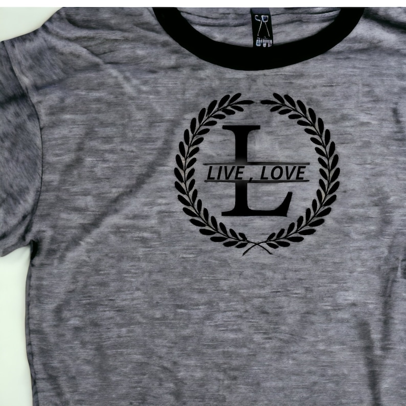 K&ouml;nnte beinhalten: Ein graues T-Shirt mit einem schwarzen gerippten Kragen. Das Shirt zeigt eine schwarze Grafik einer Lorbeerkrone mit dem Buchstaben "L" in der Mitte und dem Text "LIVE, LOVE" auf einem Banner.