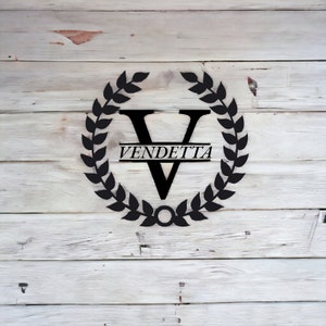K&ouml;nnte beinhalten: Schwarzes Metallmonogramm mit einem Kranz aus Bl&auml;ttern, der den Buchstaben V und das Wort "VENDETTA" auf einem wei&szlig;en Holzgrund umgibt.