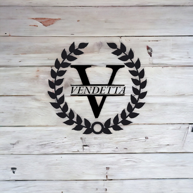 K&ouml;nnte beinhalten: Schwarzes Metallmonogramm mit einem Kranz aus Bl&auml;ttern, der den Buchstaben V und das Wort "VENDETTA" auf einem wei&szlig;en Holzgrund umgibt.